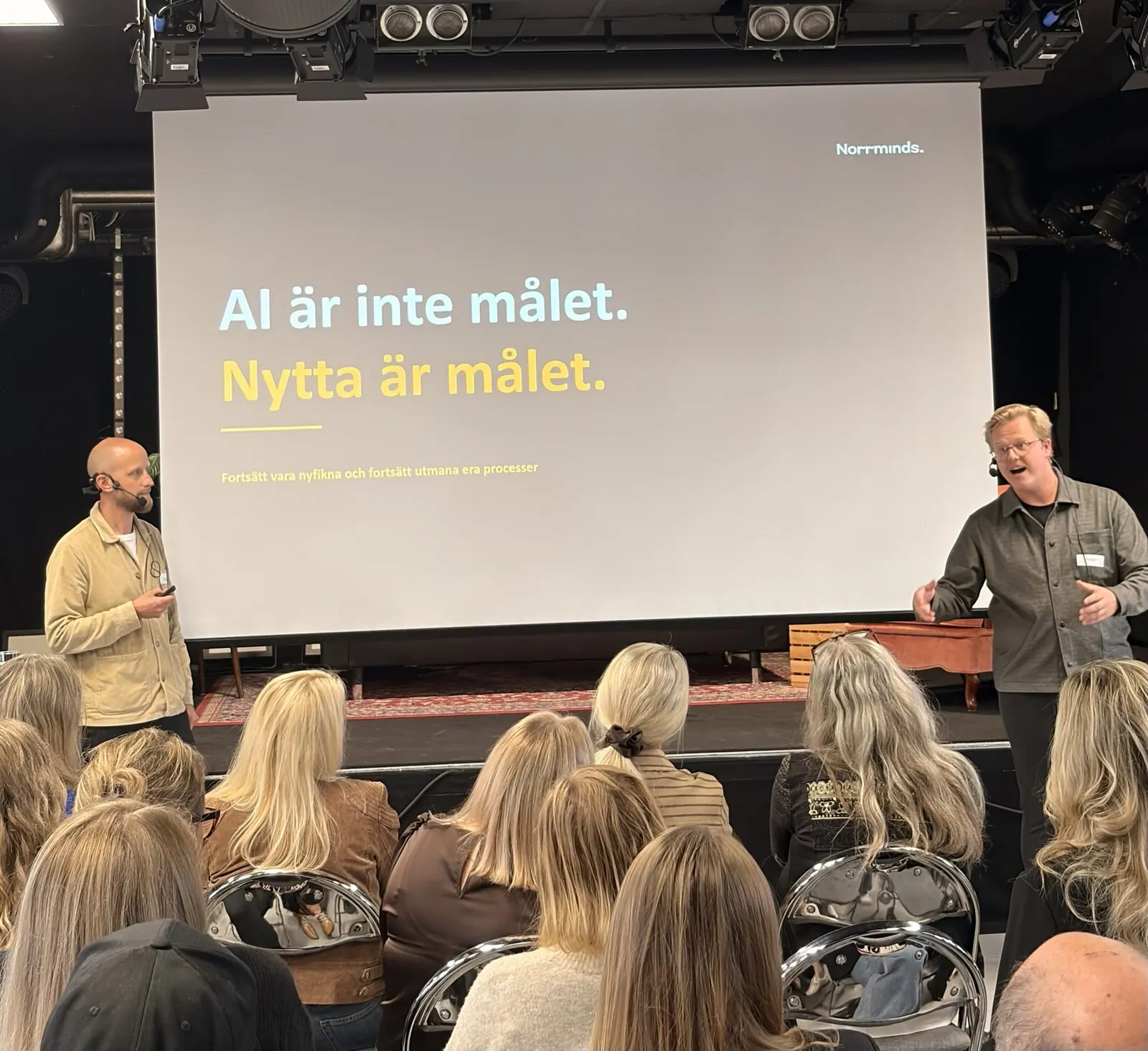 En kollega på scen bredvid en skärm med texten ”AI är inte målet. Nytta är målet.”, publiken i förgrunden.