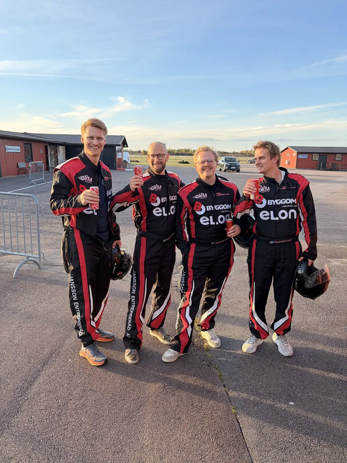 Fyra kollegor i racingoveraller skålar med läsk efter ett race på gokartbanan.
